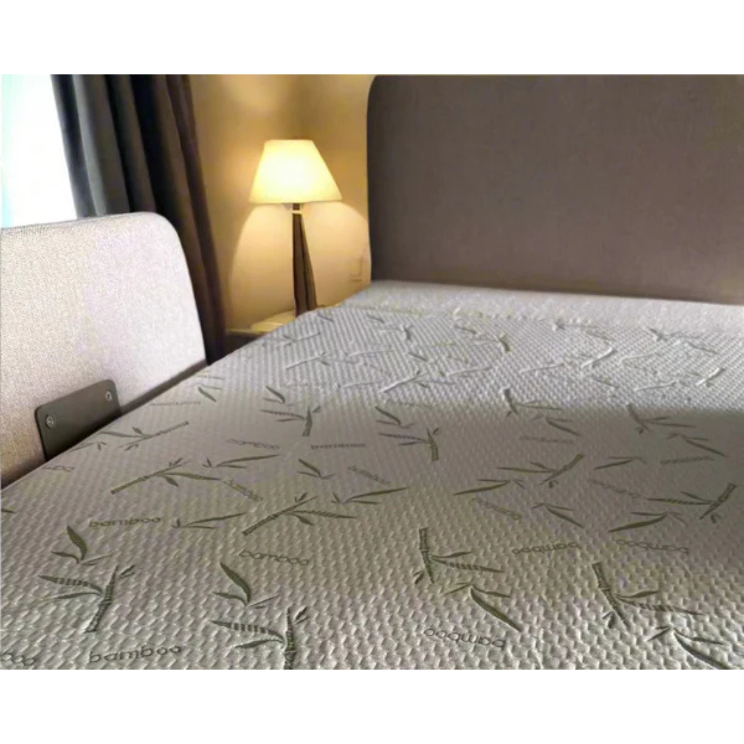 A close-up of a bed with the Waterproof Mattress Shield For Elevra Beds - PureBamboo™, showcasing its light color and subtle leaf pattern next to a beige headboard, with a lamp and curtain softly lit in the background.